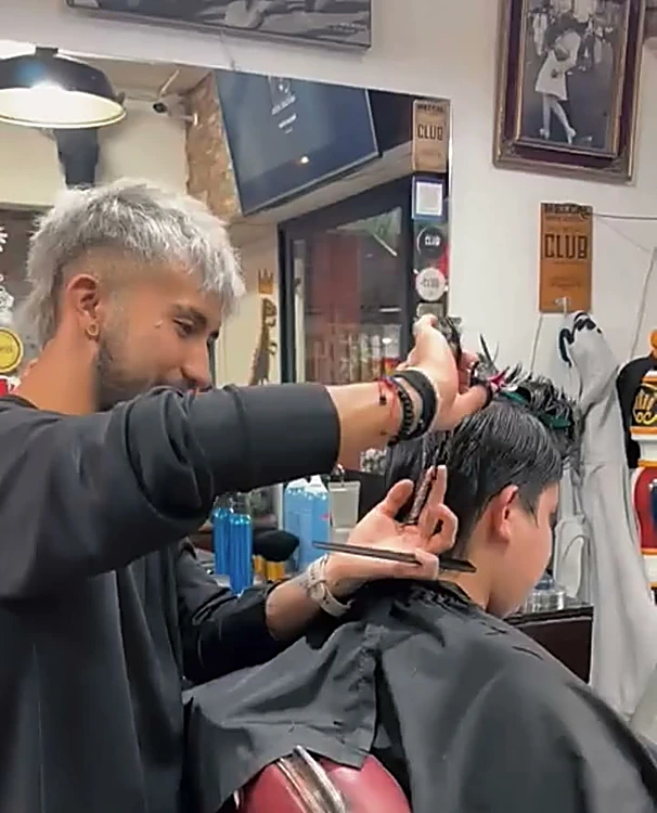 Barbero de cabello platinado cortando a tijera a un cliente de espaldas
