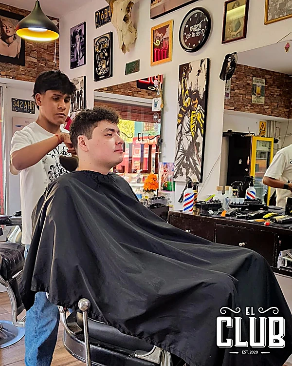 Barbero secando el cabello de un cliente frente al muro de cuadros y el poste de barbero
