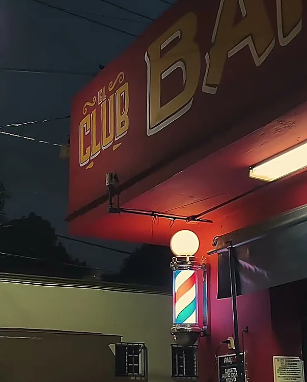 Fachada roja de El Club de noche, con el letrero EL CLUB BARBERÍA y el poste de barbero encendido