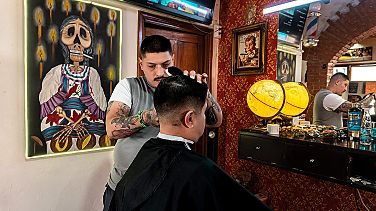 Barbero cortando junto al cuadro de una Catrina al óleo, globos terráqueos amarillos y papel tapiz vino