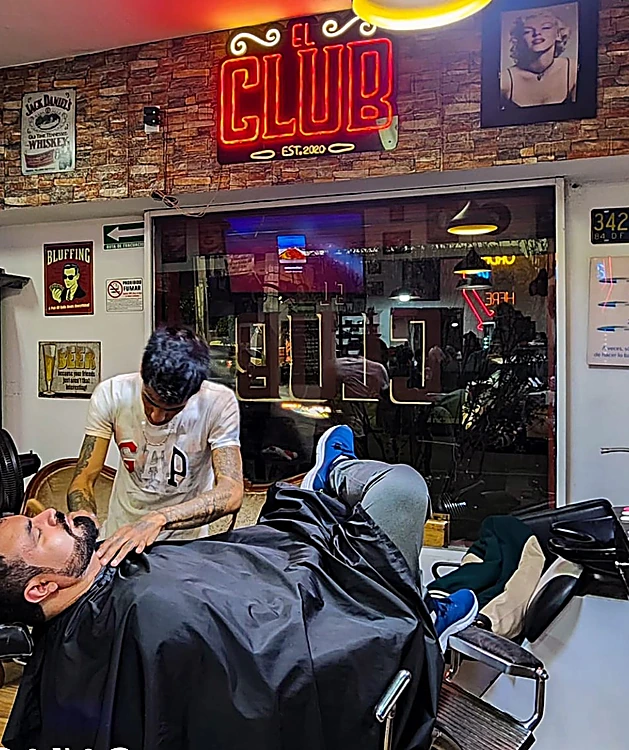 Neón rojo EL CLUB sobre el muro de ladrillo; abajo, un barbero afeita a un cliente recostado en el sillón