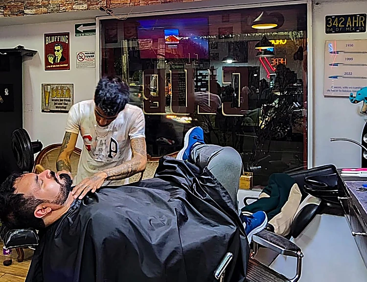 Barbero arreglando la barba de un cliente recostado bajo el neón de El Club