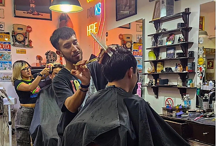 Tres barberos trabajando a la vez en el salón, con repisas de objetos de colección al fondo