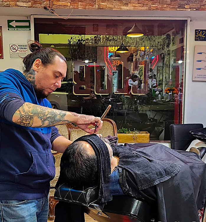 Barbero tatuado afeita con navaja a un cliente recostado con la toalla sobre el pecho